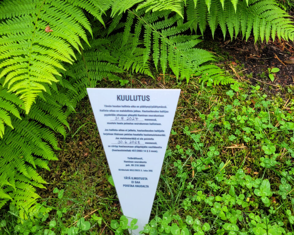 Kuulutuslappu haudalla saniaisten edessä.